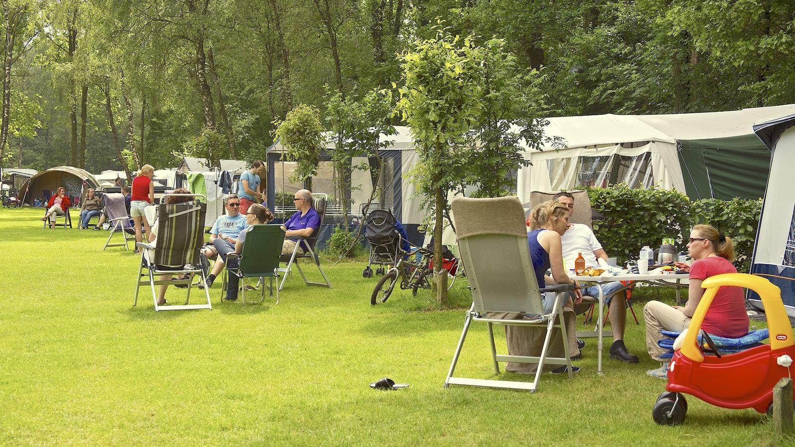 Gasten zitten in kampeerstoelen rond een tafel voor hun caravan, met kinderen die op het gras spelen